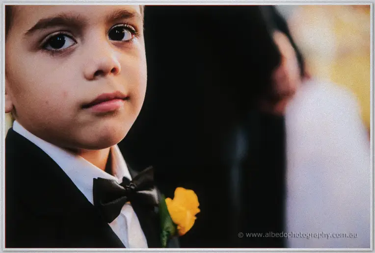 A boy in a tuxedo and bow tie watching the ceremony with a yellow rose pinned to his lapel