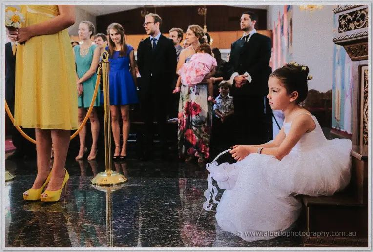 A little flower girl in white sitting alone on the church steps while guests stand watching nearby