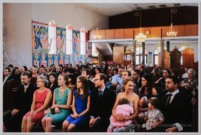 Wedding congregation seated in wooden pews beneath bright icon murals and hanging chandeliers