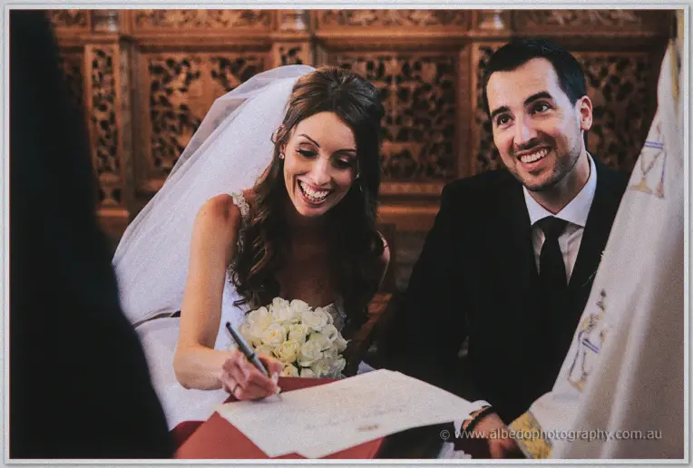 Bride laughing as she signs the marriage register while her groom grins broadly beside her in the church