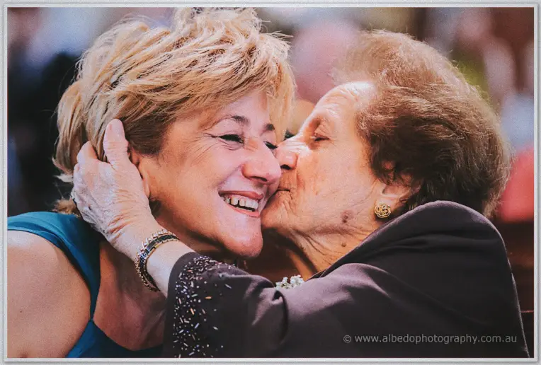 An older guest kisses a laughing woman in teal on the cheek at Michele and Nick's church ceremony.