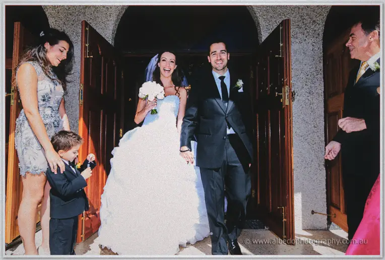 Michele and Nick walk beaming from the church doors into sunlight with their page boy and guests close behind.