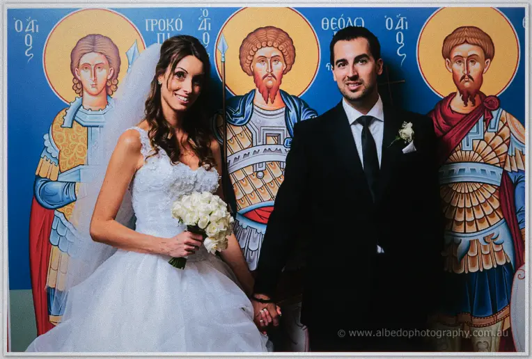 Michele and Nick hold hands before a blue wall of Byzantine warrior-saint icons, smiling warmly.