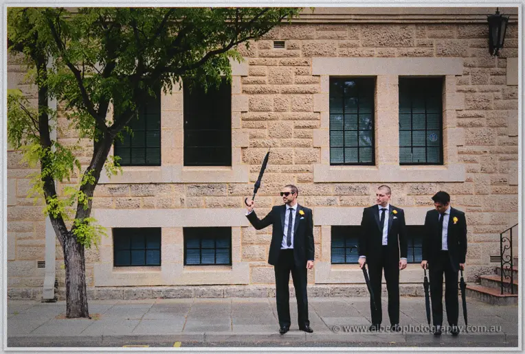 Groomsmen twirl black umbrellas on the footpath by a honey-toned stone wall before the ceremony.