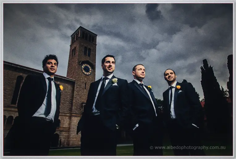 From below, Nick and his groomsmen loom under heavy grey clouds beside the sandstone clock tower.