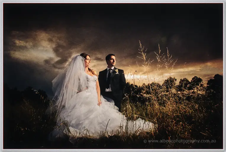 Bride in a long veil and groom in a black suit standing in tall grass beneath a stormy sunset sky