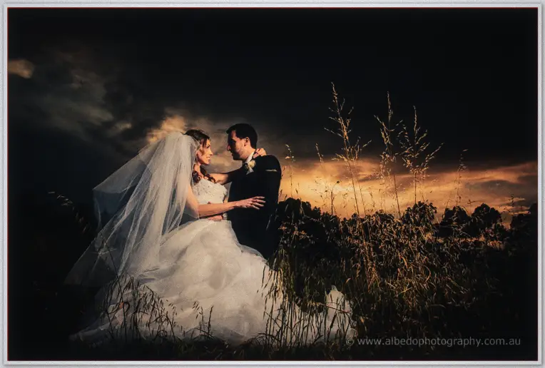 Michele and Nick embrace in tall golden grass beneath a fiery sunset sky after the storm.