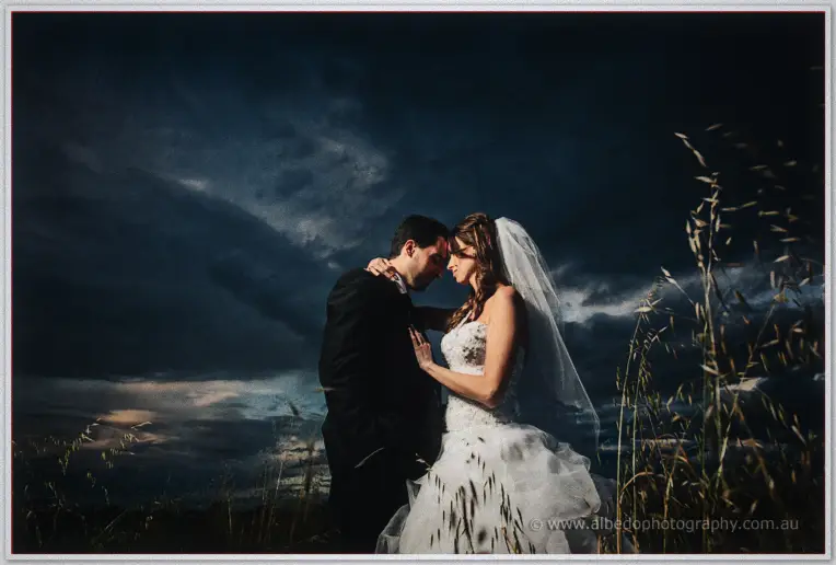 Forehead to forehead, the couple stands close in the windswept grass under a moody evening sky.