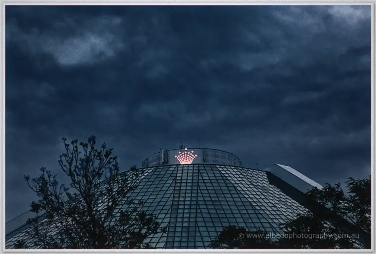 The glowing dome of Crown Perth rises into a stormy dusk sky above the trees.