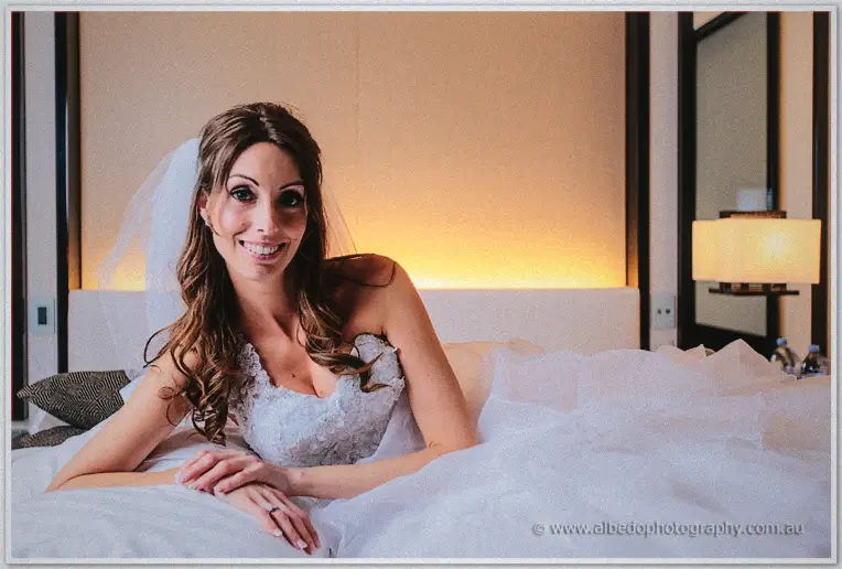 Michele reclines on the white hotel bed, veil spread, smiling in the lamplight.
