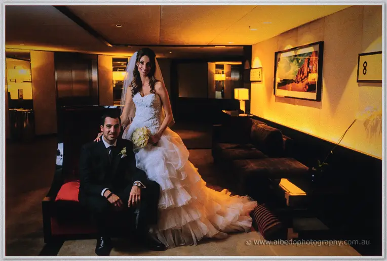 Nick sits on a red lounge as Michele rests a hand on his shoulder in the gold-lit hotel lobby.