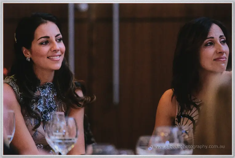 Two guests in embellished dresses smile during speeches at the candlelit ballroom table.