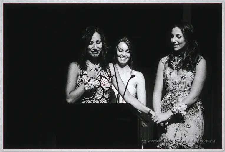 Bridesmaids clasp hands at the podium, one wiping away tears mid-wedding speech.