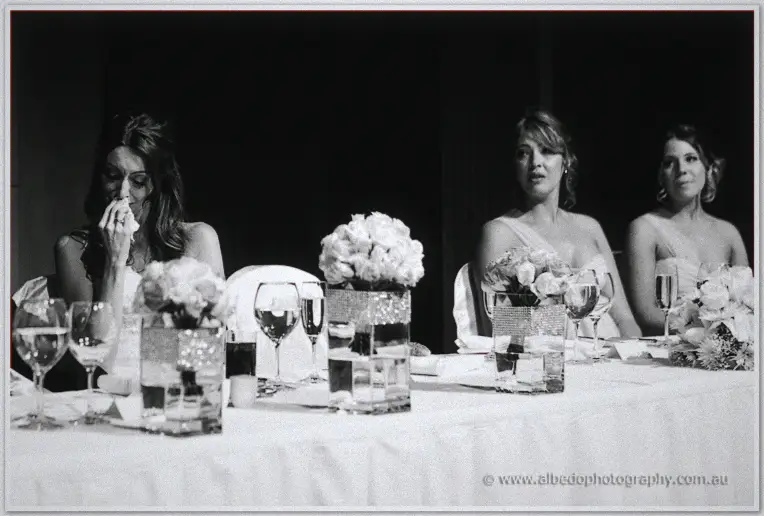 A bridesmaid dabs tears at the bridal table as speeches unfold under soft monochrome light.