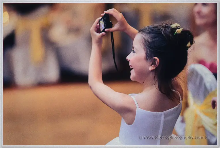 A little flower girl in white aims a compact camera across the yellow-sashed ballroom.