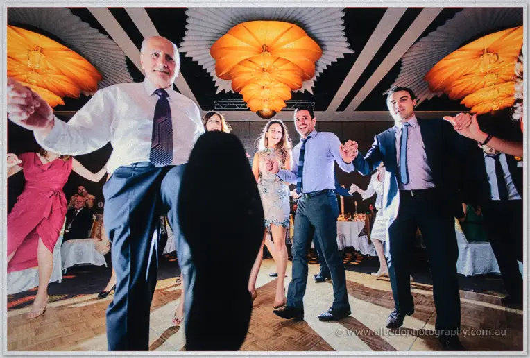 Guests kick high in a hand-in-hand Greek circle dance across the ballroom parquetry.