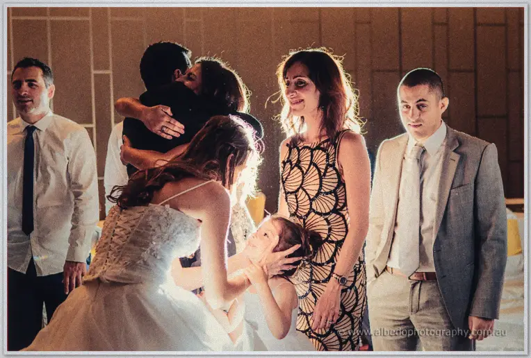 Michele crouches to a little girl's level as her parents embrace in the warm reception glow.