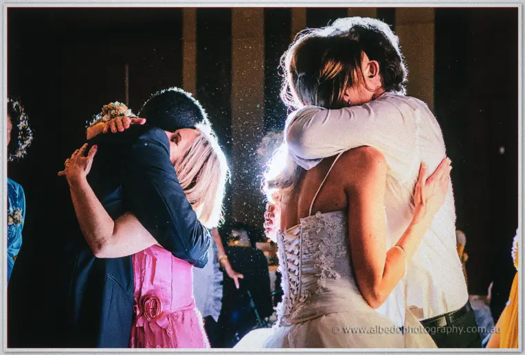 Bride in a strapless lace gown embracing a guest as couples hug on the warmly lit Astral Ballroom dance floor