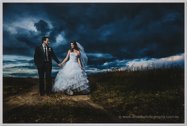 Michele and Nick hold hands walking a dirt track through open grassland beneath dramatic storm clouds at dusk
