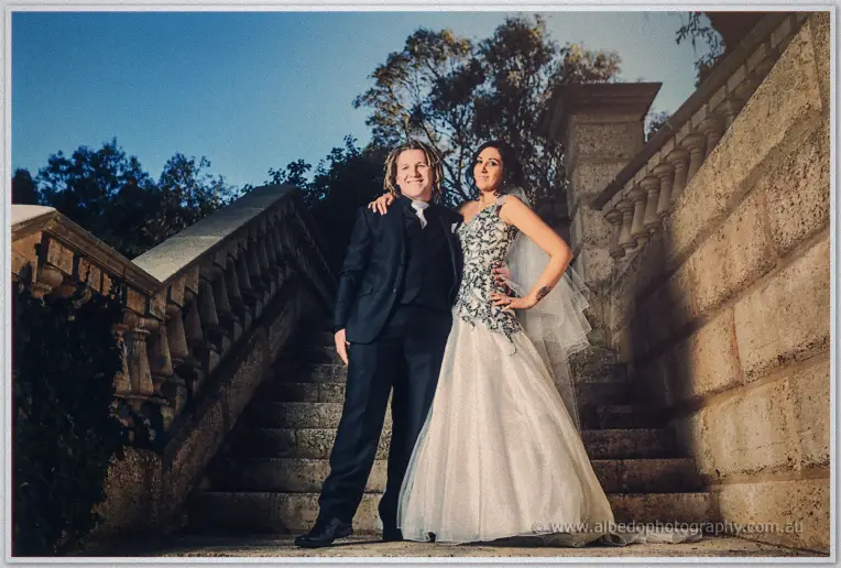 Jenna rests her hand on Andrew's shoulder on stone steps at dusk, her gown and veil catching the fading light.