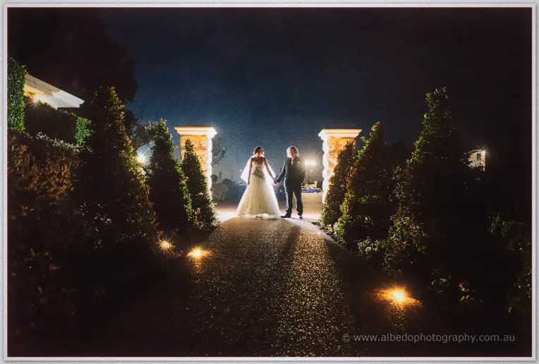 Jenna and Andrew stand hand in hand between floodlit stone pillars on the garden path at night.