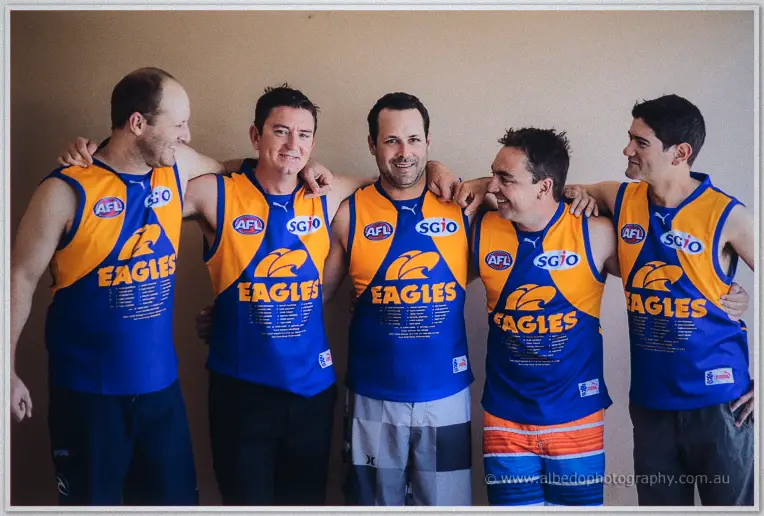Five groomsmen arm in arm in blue and gold Eagles football jumpers, laughing against a plain wall