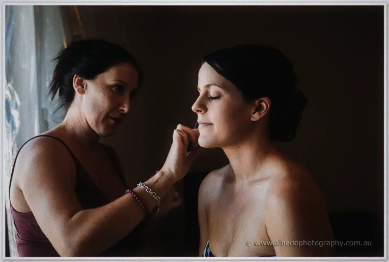 Makeup artist applying cosmetics to bride Allison by soft window light as she closes her eyes