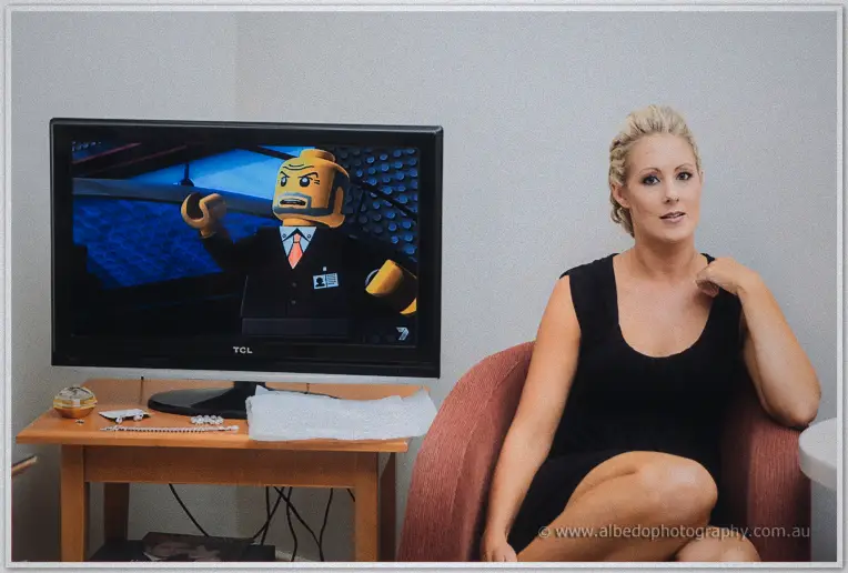 Bridesmaid in a black dress seated in an armchair beside a TV playing a Lego figure cartoon