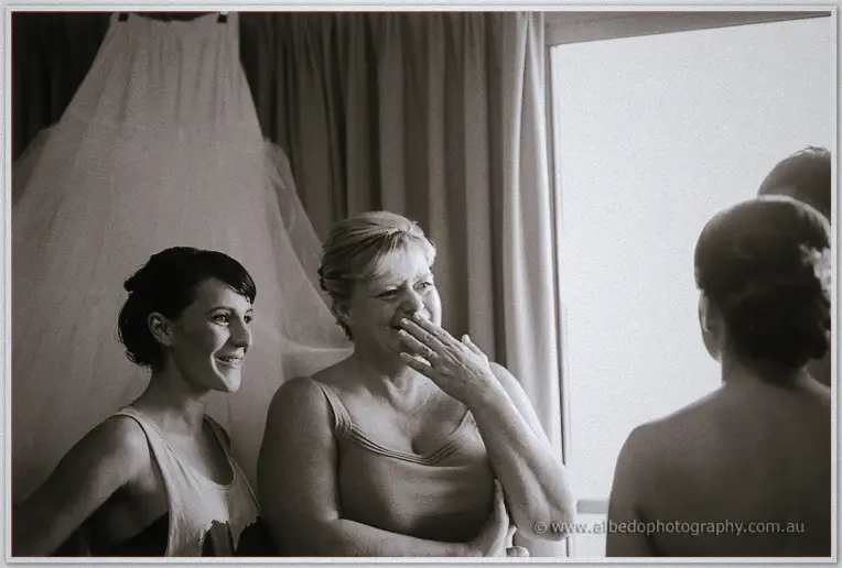 Bridesmaid and mother laughing near the hanging wedding dress as Allison has her final touches done