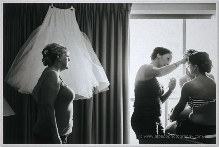 Black and white scene of the wedding dress hanging by a curtained window while makeup is finished