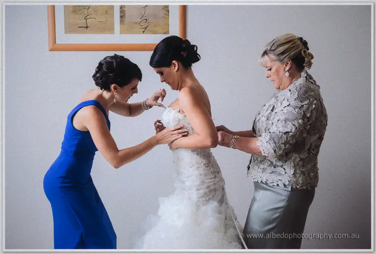 Bridesmaid and mother fastening Allison into her lace-detailed gown in bright daylight