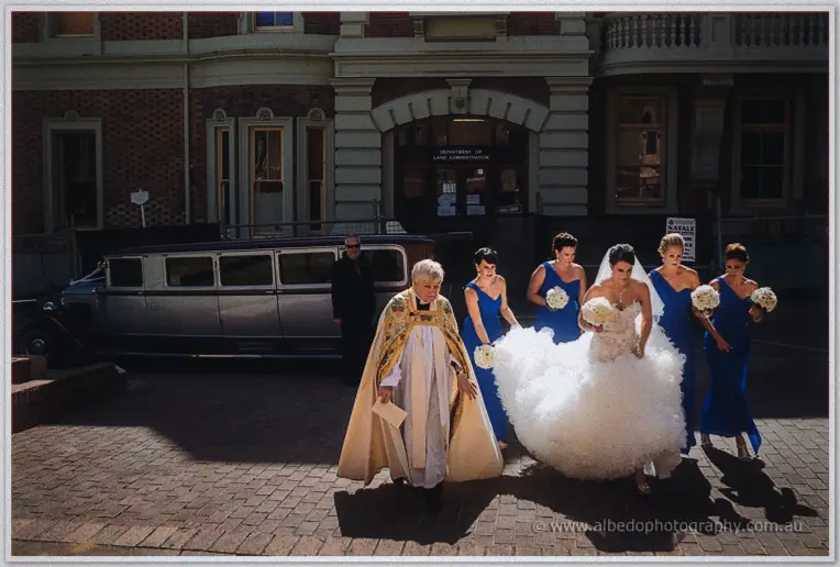 Priest in gold vestments greets Allison and her bridal party arriving outside St George's Cathedral