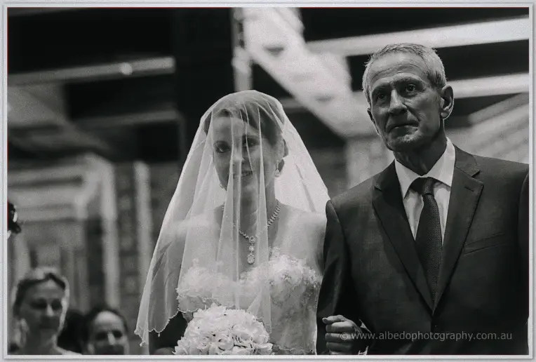 Black and white portrait of a veiled Allison beside her father as they reach the cathedral altar