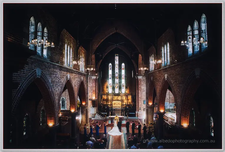 High wide view of the bride and groom at the altar inside the brick cathedral beneath glowing stained glass