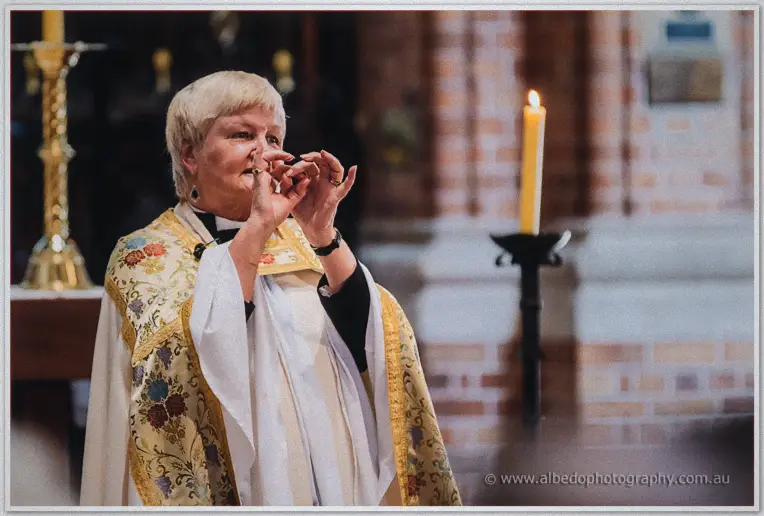 Priest in embroidered vestments gesturing during the vows beside tall cathedral candles