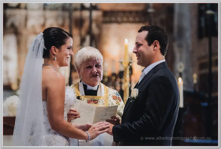 Allison and Greg holding hands and smiling at each other as the priest reads their vows