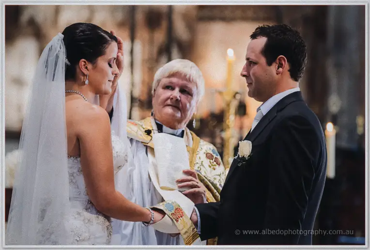 Veiled Allison and Greg holding hands before the priest during their wedding vows