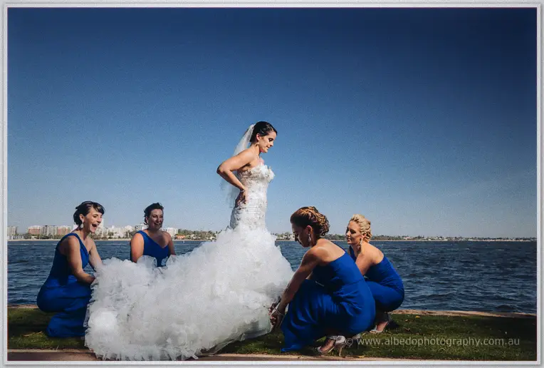 Bridesmaids in blue arranging Allison's ruffled train on the riverfront grass under a clear sky