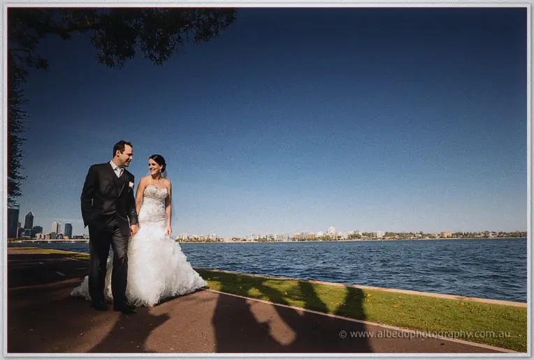 Allison and Greg strolling hand in hand along the river path with the Perth skyline behind
