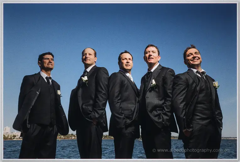 Greg and his groomsmen in black suits gazing upward against a deep blue riverside sky