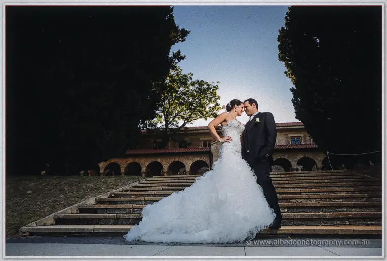Allison and Greg on sunlit stone steps beneath an arched colonnade, her ruffled train spread wide.