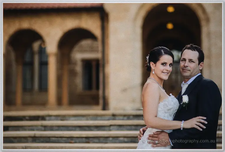 Allison and Greg smiling into each other's eyes on cathedral steps, sandstone arches soft behind them.