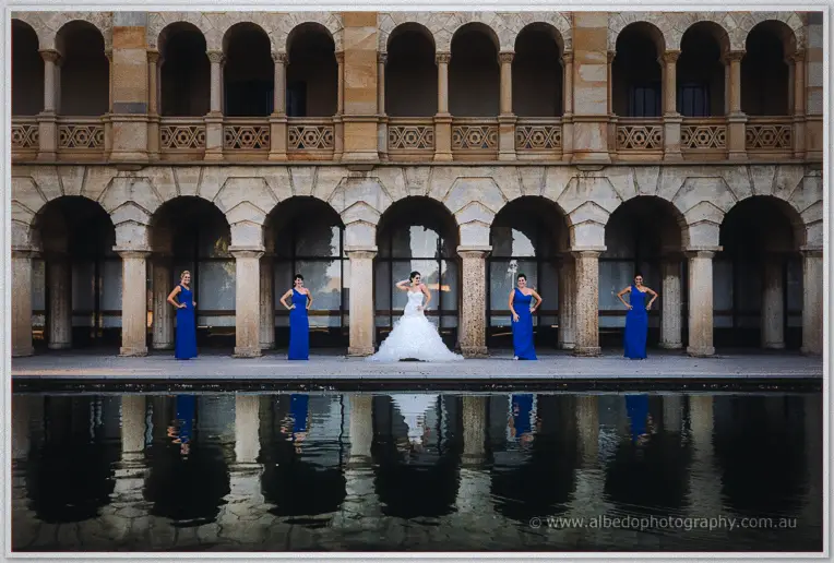 Allison and her four bridesmaids in cobalt gowns striking poses along a colonnade mirrored in dark water.