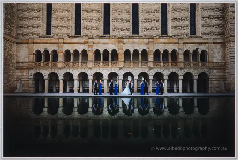 Bridal party in blue gowns and dark suits lined up along a sandstone colonnade, mirrored in still water.