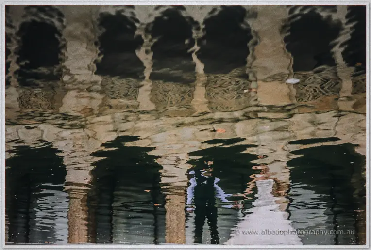 The couple's reflections wobble across dark pool water, the colonnade melting into abstract ripples above.