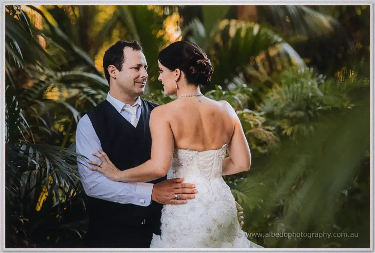 Greg smiling at Allison across her bare shoulder, her lace-back gown and necklace picked out in warm light.