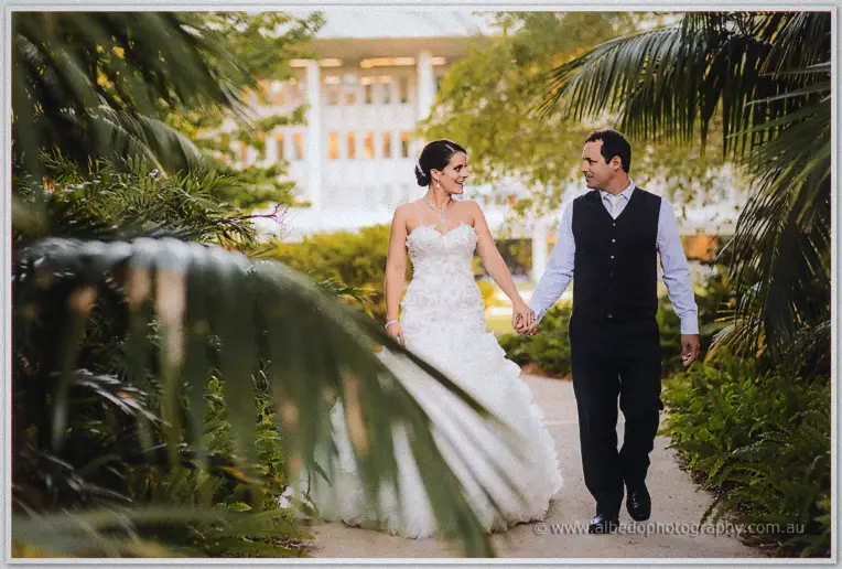 Hand in hand down a palm-lined garden path, the white facade of Frasers glowing softly in the distance.