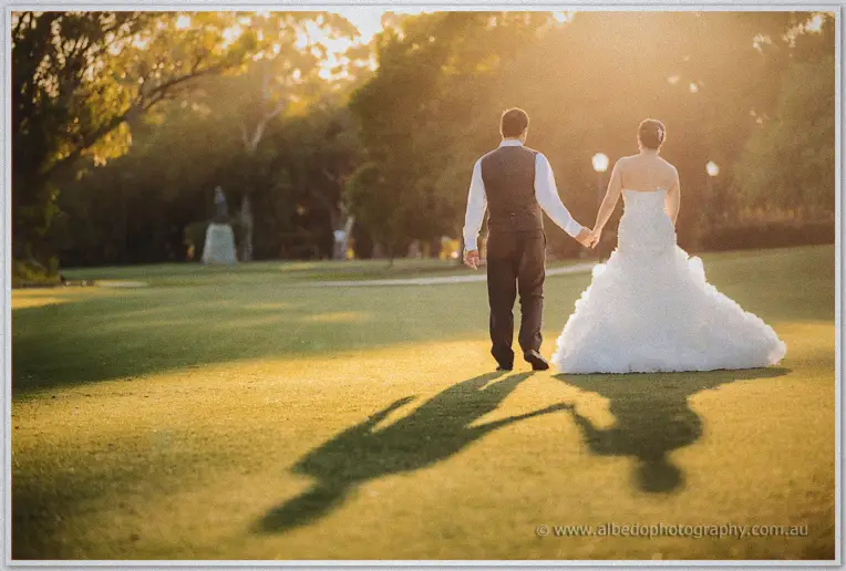 Allison and Greg walk hand in hand across sunlit lawns at golden hour, long shadows on the grass