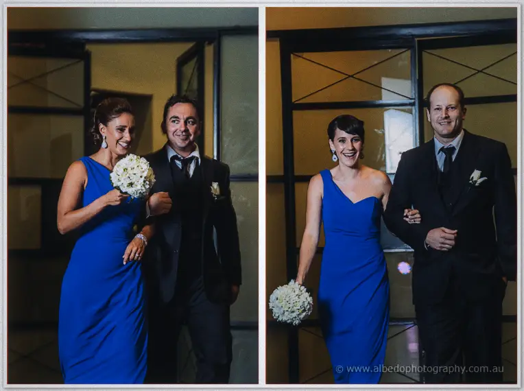 Bridesmaids in royal blue enter arm in arm with groomsmen, clutching white hydrangea posies and grinning.