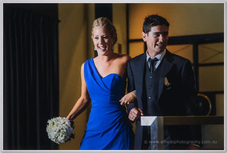 Another laughing bridesmaid in a one-shoulder cobalt gown walks in beside a beaming groomsman.